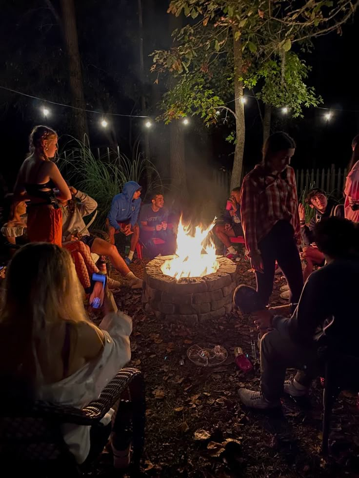 a group of people sitting around a fire pit in the woods at night with lights on