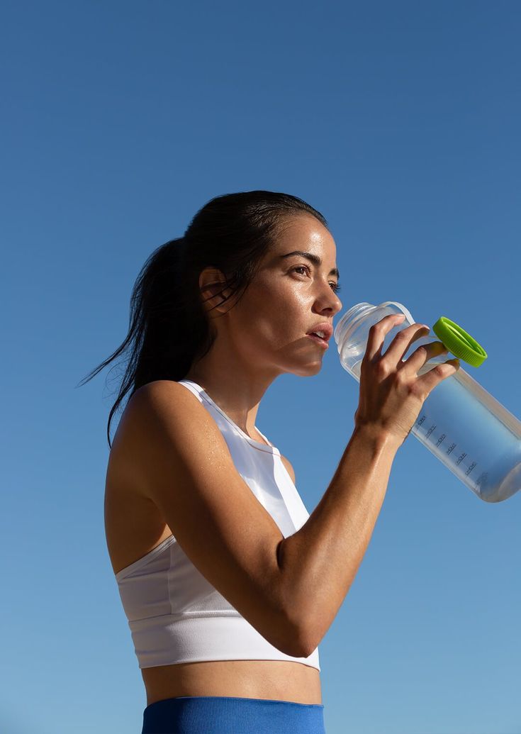 a woman drinking from a water bottle while standing in front of a blue cloudy sky