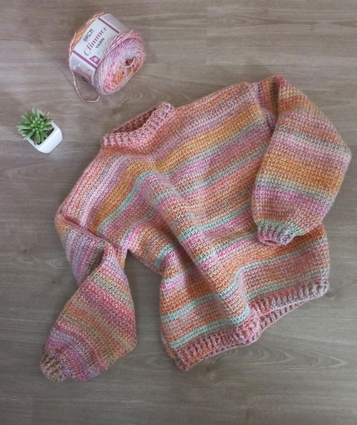 a knitted sweater next to a potted plant on a wooden floor with a spool of thread