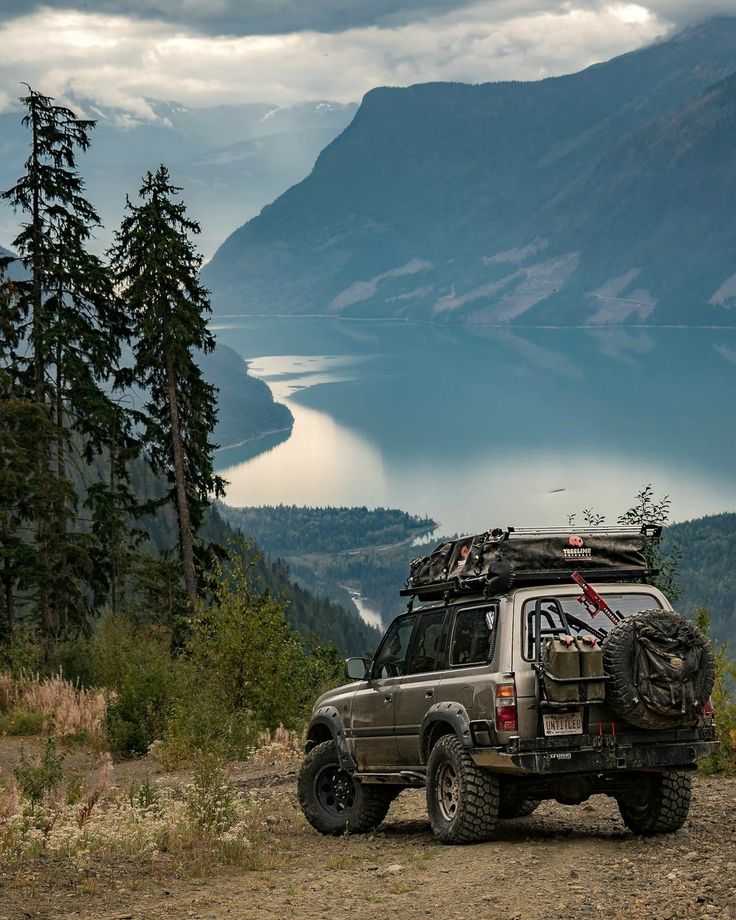 an suv parked on the side of a mountain with a lake and mountains in the background