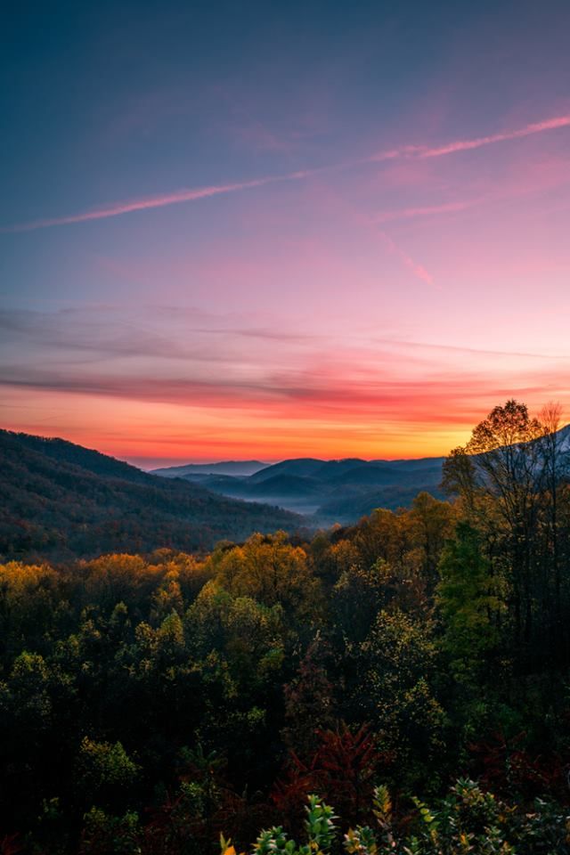 the sun is setting over mountains and trees in the foreground, as seen from an overlook