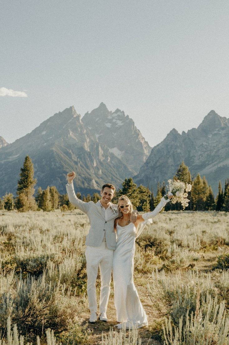 a newly married couple standing in the middle of a field with mountains in the background