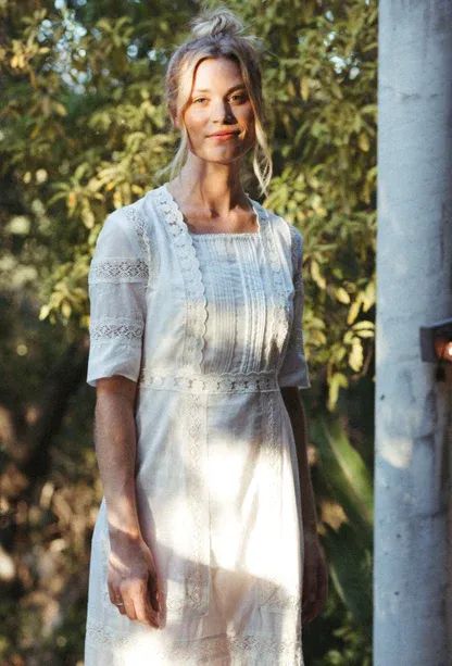 an older woman in white dress standing next to a pole with trees in the background