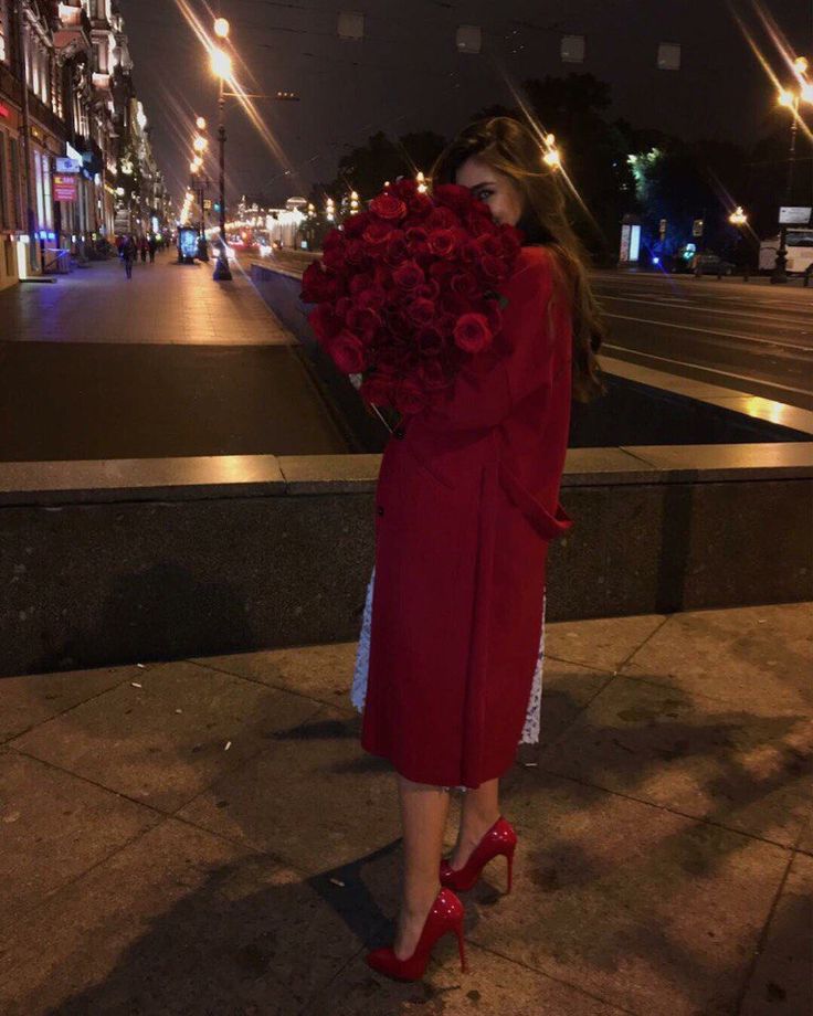 a woman in a red coat is standing on the sidewalk with roses around her neck