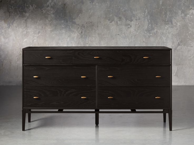 a black dresser with gold knobs on the top and bottom drawers, in front of a concrete wall