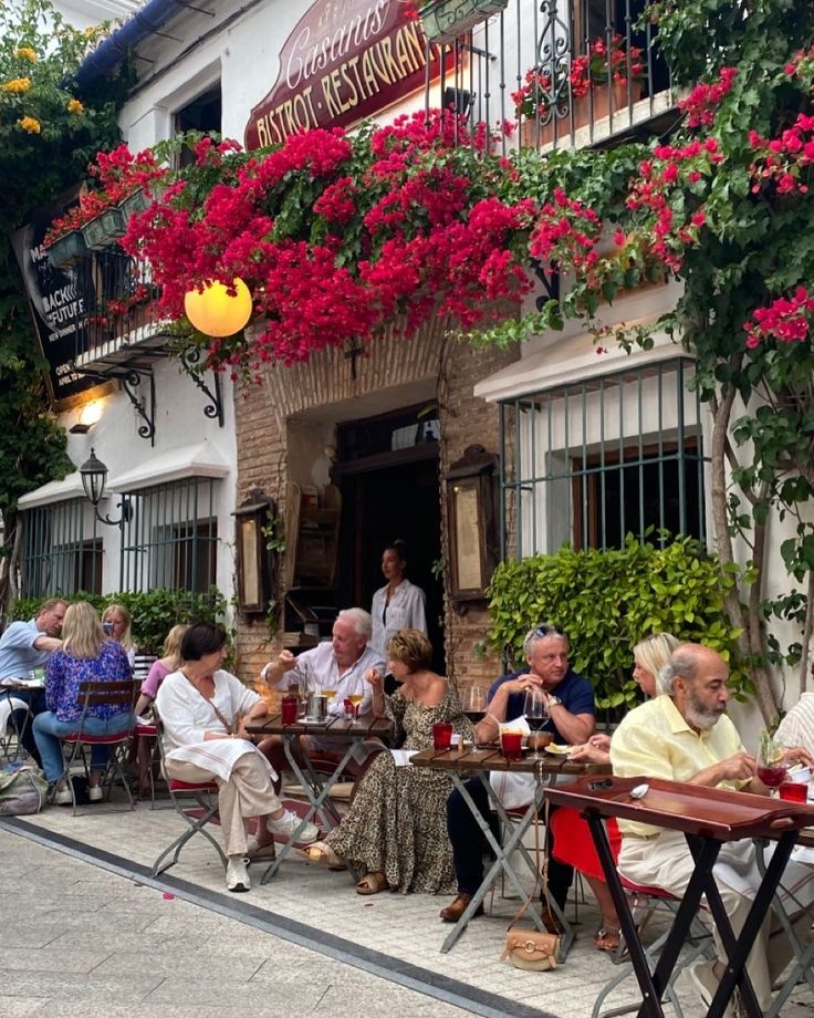 many people are sitting at tables in front of a building with red flowers on it