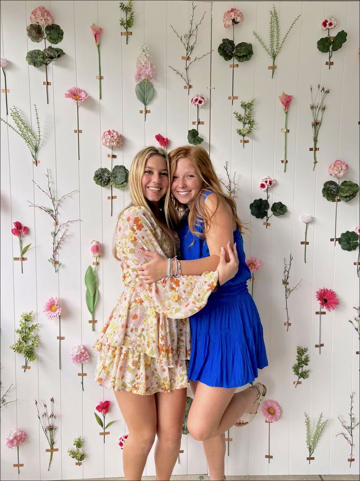 two young women hugging each other in front of a wall with flowers and plants on it