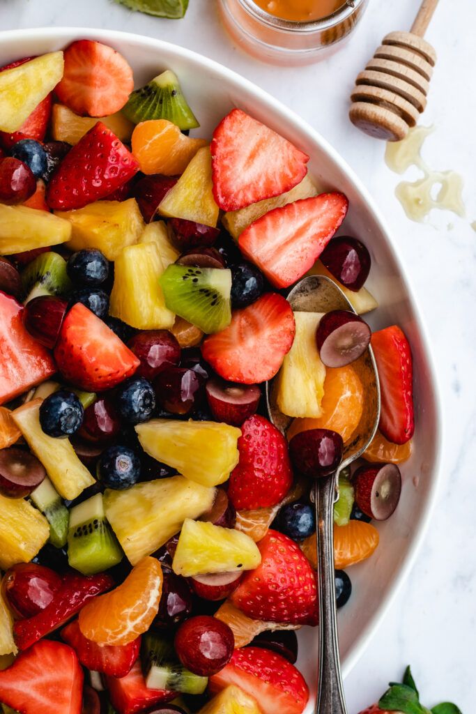 a white bowl filled with fruit salad next to a jar of honey and spoons