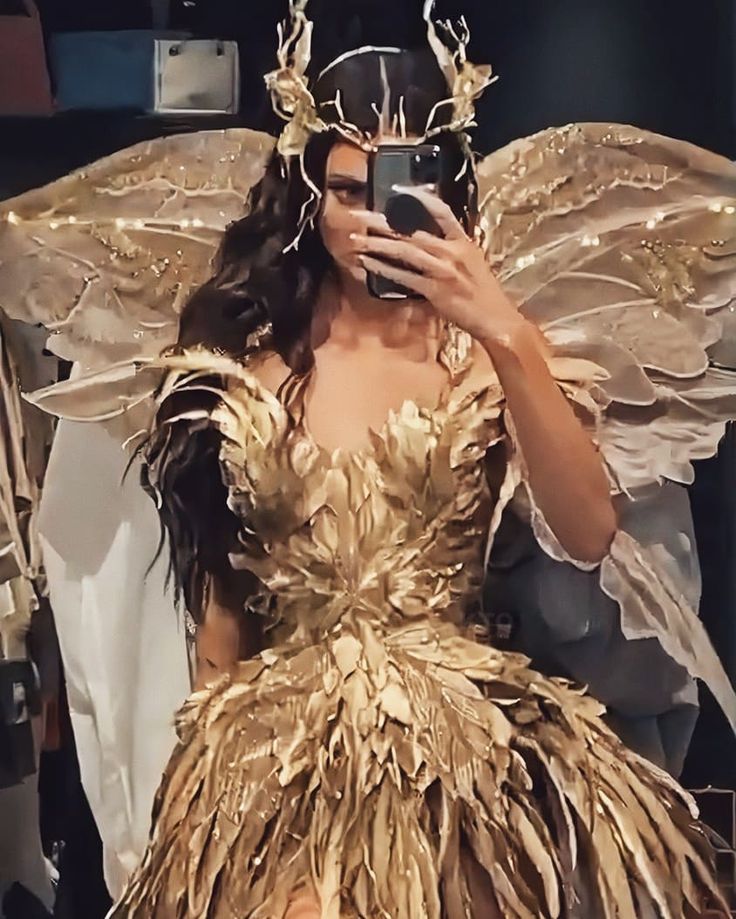 a woman in a gold dress takes a selfie with her cell phone while she is dressed as a fairy