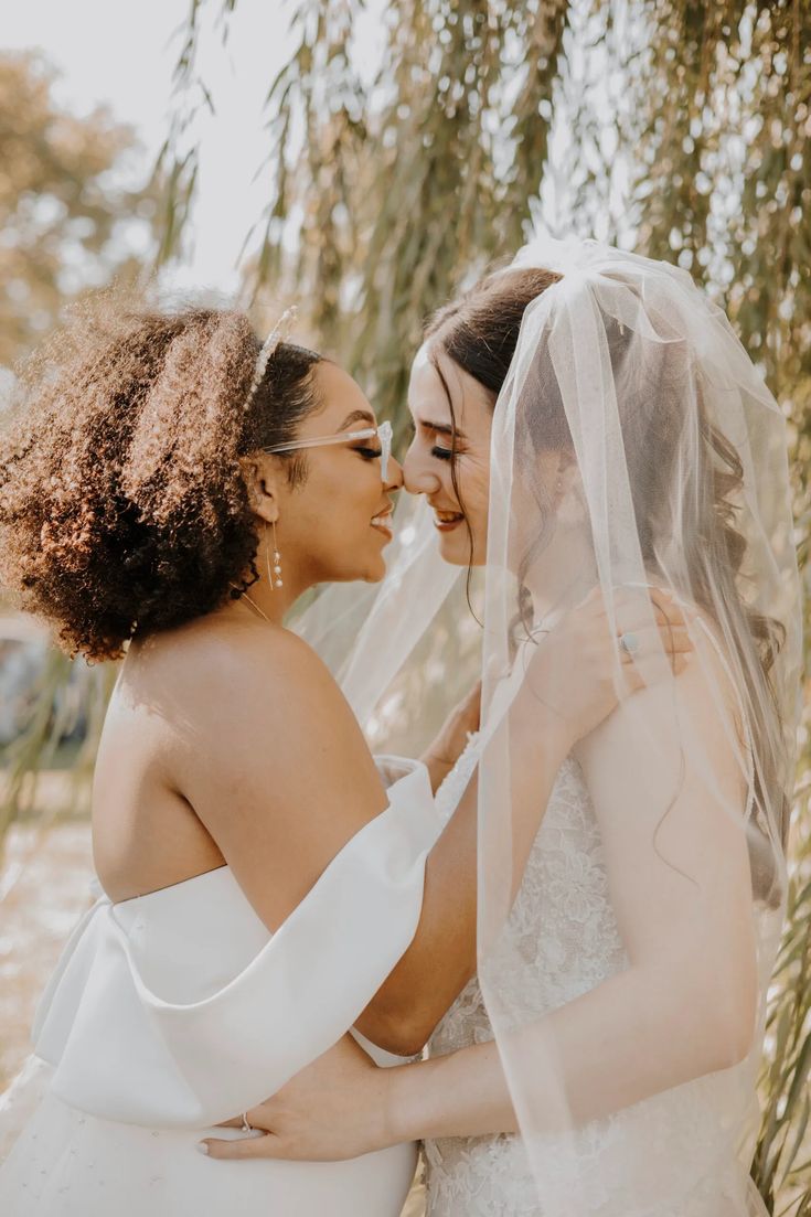 two women in wedding gowns standing next to each other and one is kissing the other