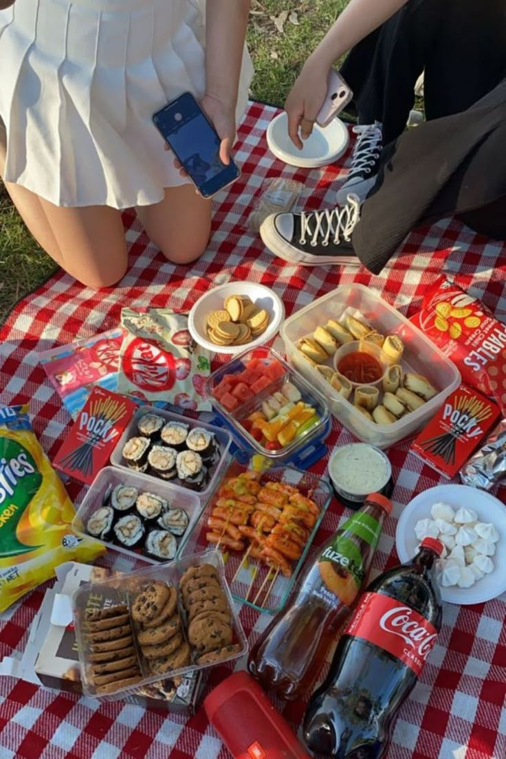 two people sitting at a picnic table with food and drinks on it, one person is holding a cell phone