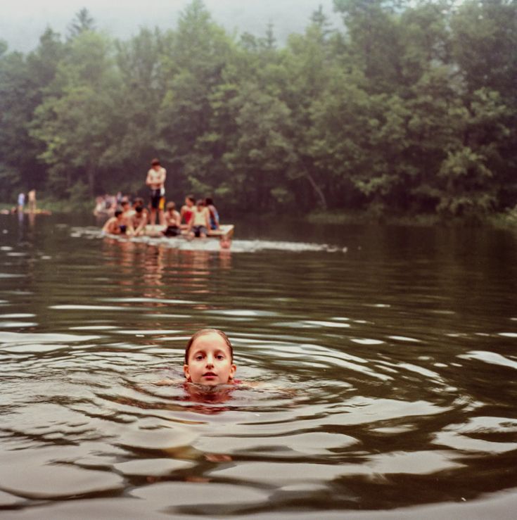 a woman swimming in the water with people floating on boats behind her and trees in the background