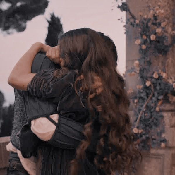 a woman with long curly hair hugging another woman's back in front of an old building