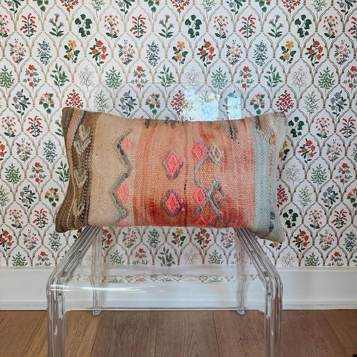 a decorative pillow sitting on top of a clear bench in front of a wallpapered background