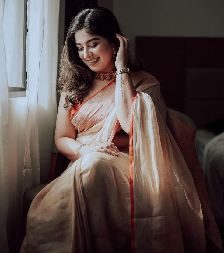 a woman sitting on top of a bed next to a window wearing a white and orange sari