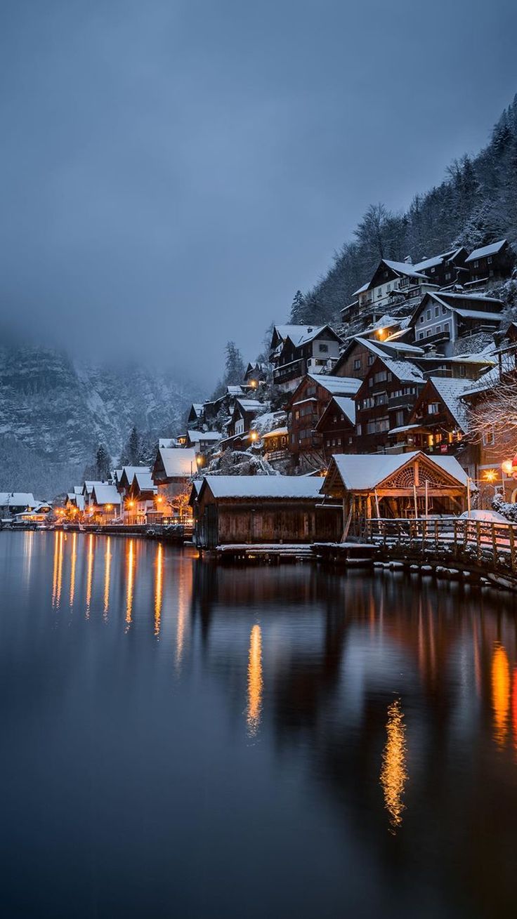 a lake with snow covered buildings and lights on the water at night in front of mountains