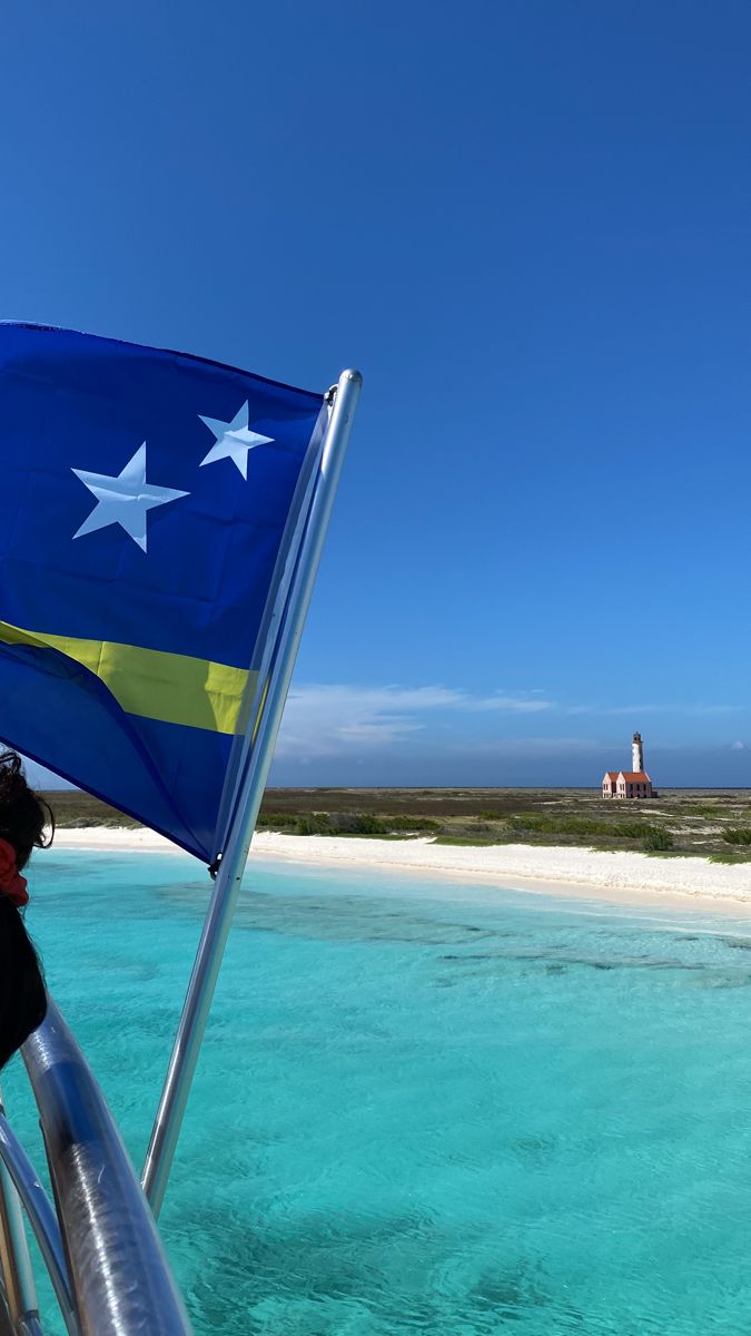 a flag flying on the back of a boat in clear blue water with an island in the background