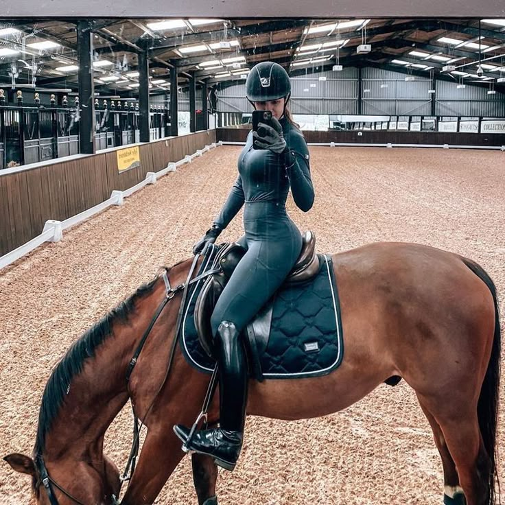 a woman riding on the back of a brown horse in an indoor arena while talking on a cell phone