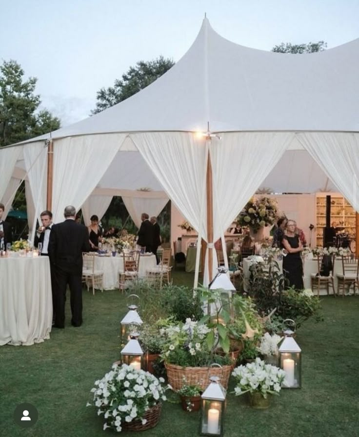 a group of people standing under a white tent next to tables and chairs with flowers on them