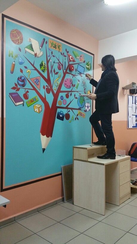 a man standing on top of a wooden desk in front of a tree with school supplies painted on it