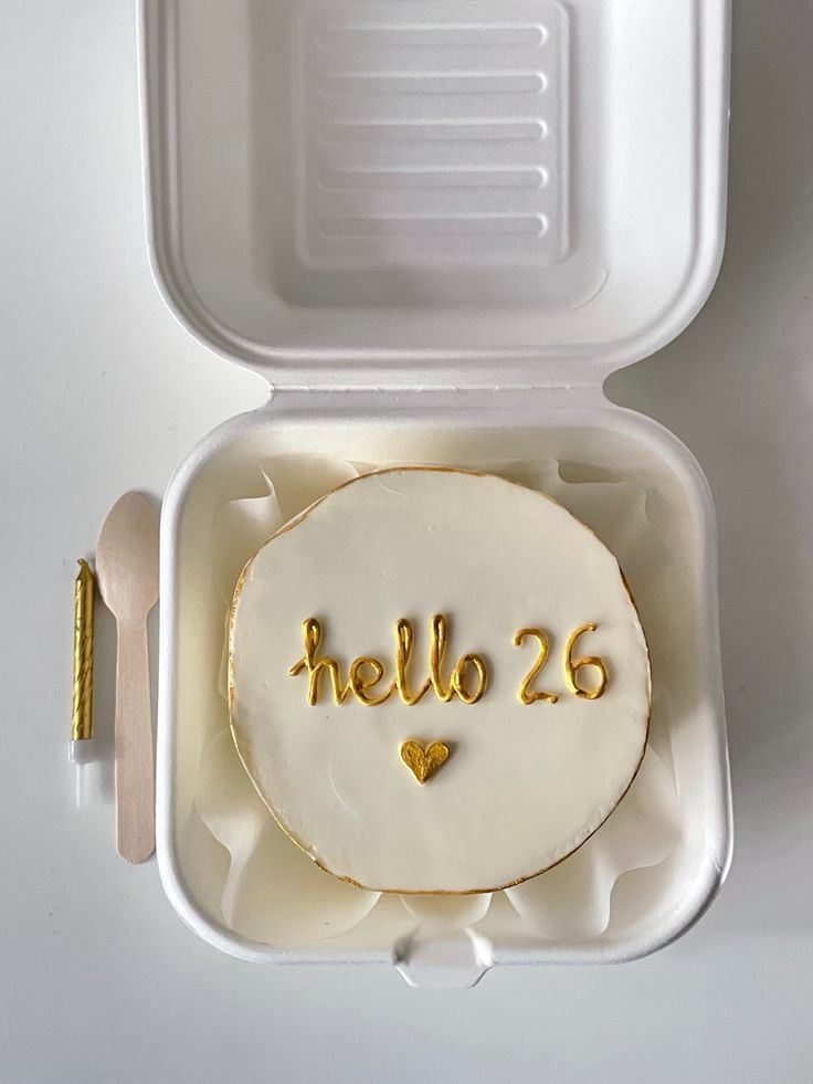 a hello 26 cake in a white plastic container with gold lettering on it and a fork