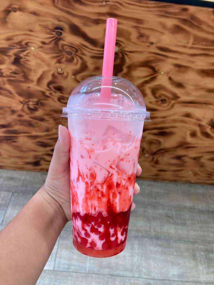 a hand holding a plastic cup with red liquid and strawberries on the rim, in front of a wooden wall