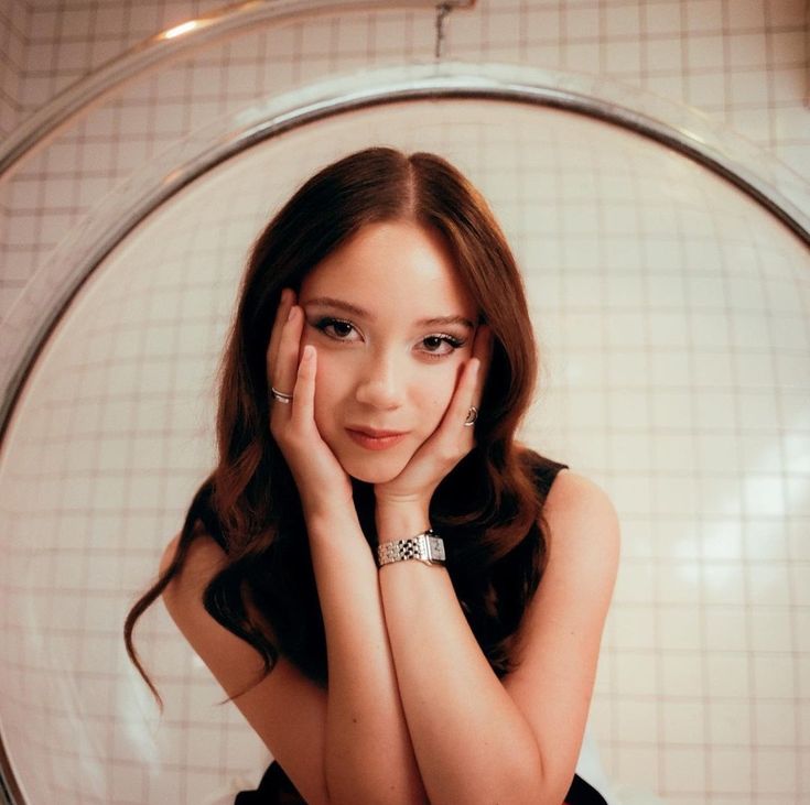 a young woman sitting in front of a mirror with her hands on her face and looking at the camera