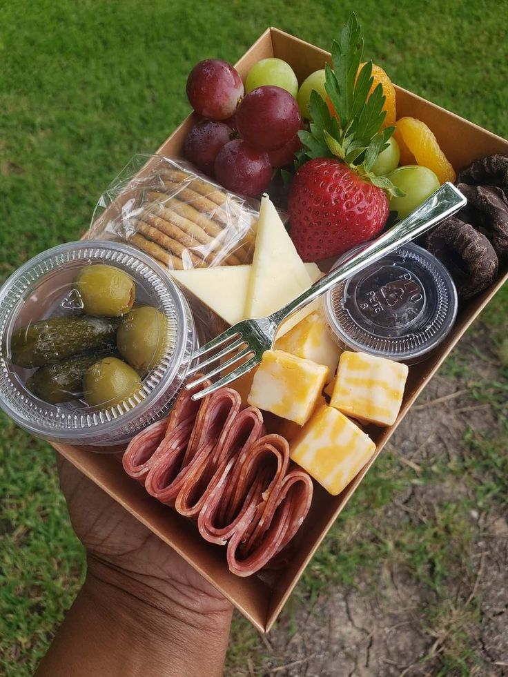 a person holding a box full of cheese, crackers, fruit and sausages