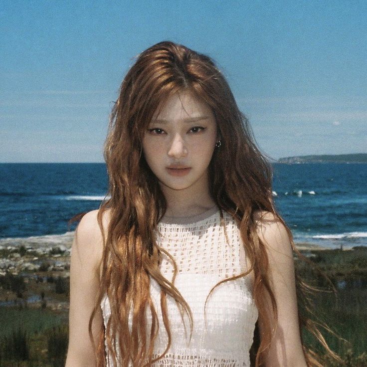 a young woman with long hair standing in front of the ocean and looking at the camera