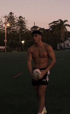 a shirtless man holding a rugby ball on a field at dusk with trees in the background