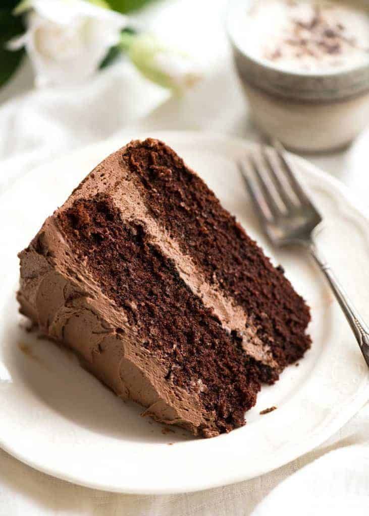 a slice of chocolate cake on a white plate with a fork and cup in the background