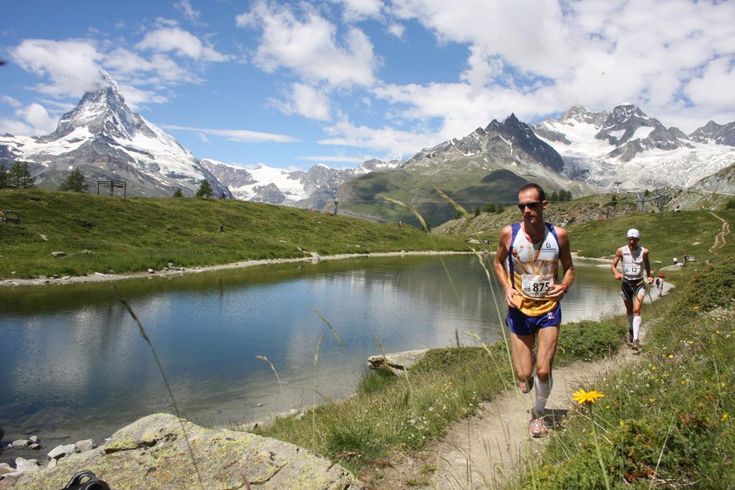 two people are running on a trail near a lake with mountains in the back ground