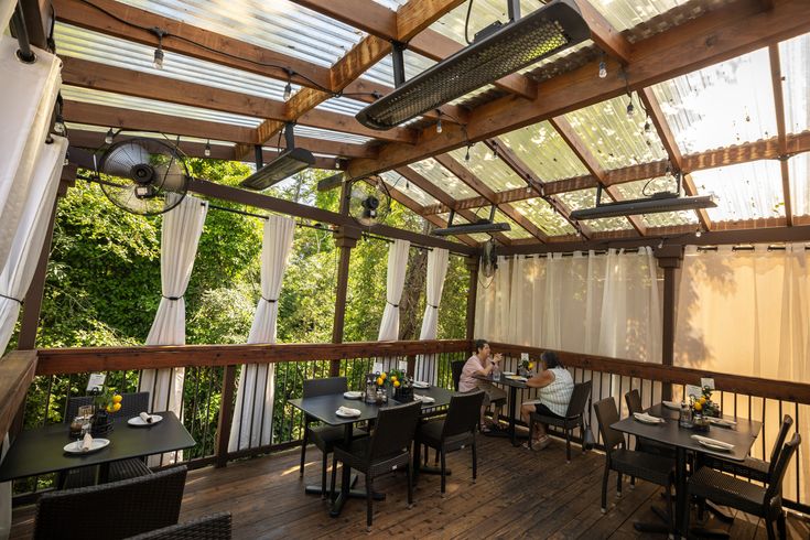 people are sitting at tables in an outdoor dining area with sheer curtains on the windows