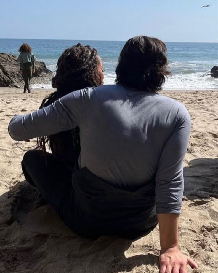 two people are sitting on the sand at the beach