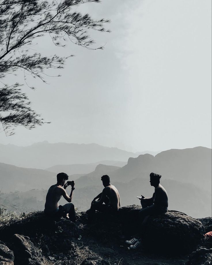 three people sitting on top of a mountain with a tree in the foreground and mountains in the background