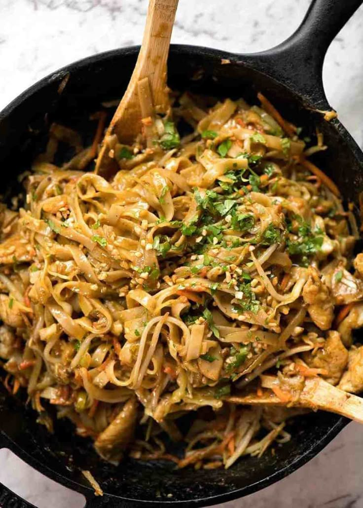 a skillet filled with noodles and vegetables on top of a marble countertop next to a wooden spoon