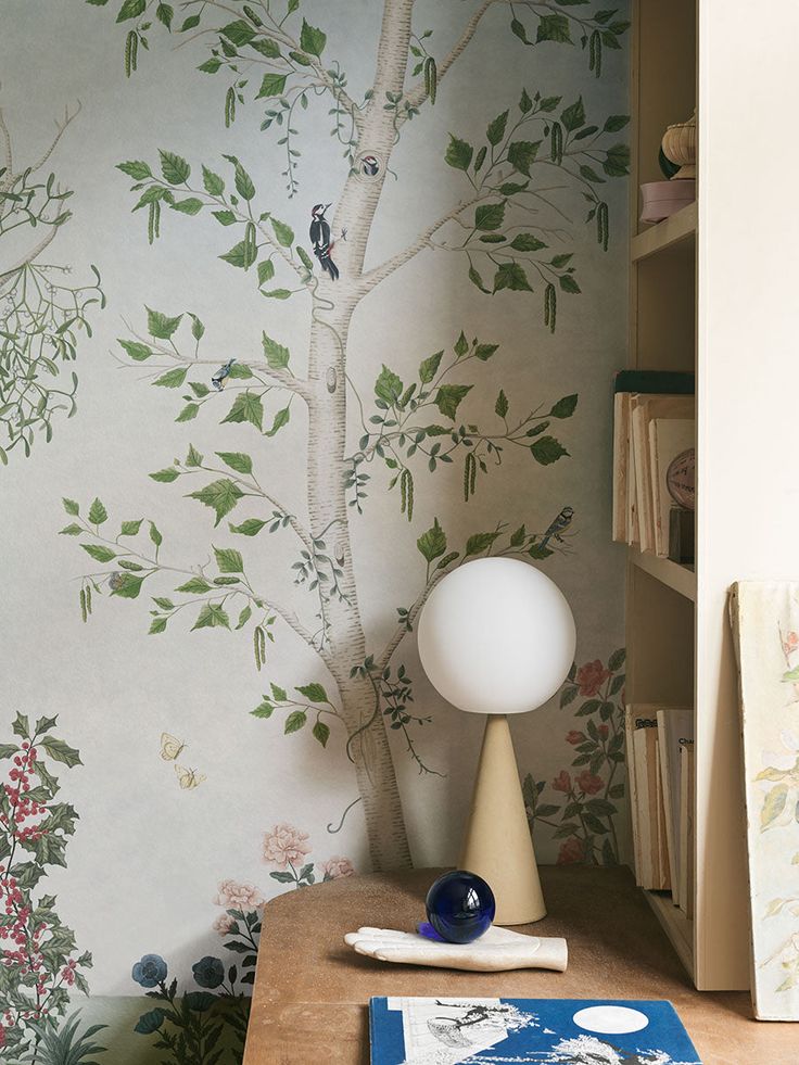 a desk with a lamp on it next to a wallpapered tree and bookshelf