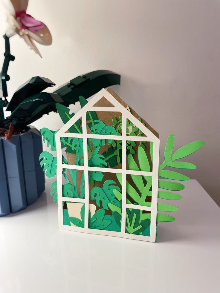 a house made out of paper sitting next to a potted plant