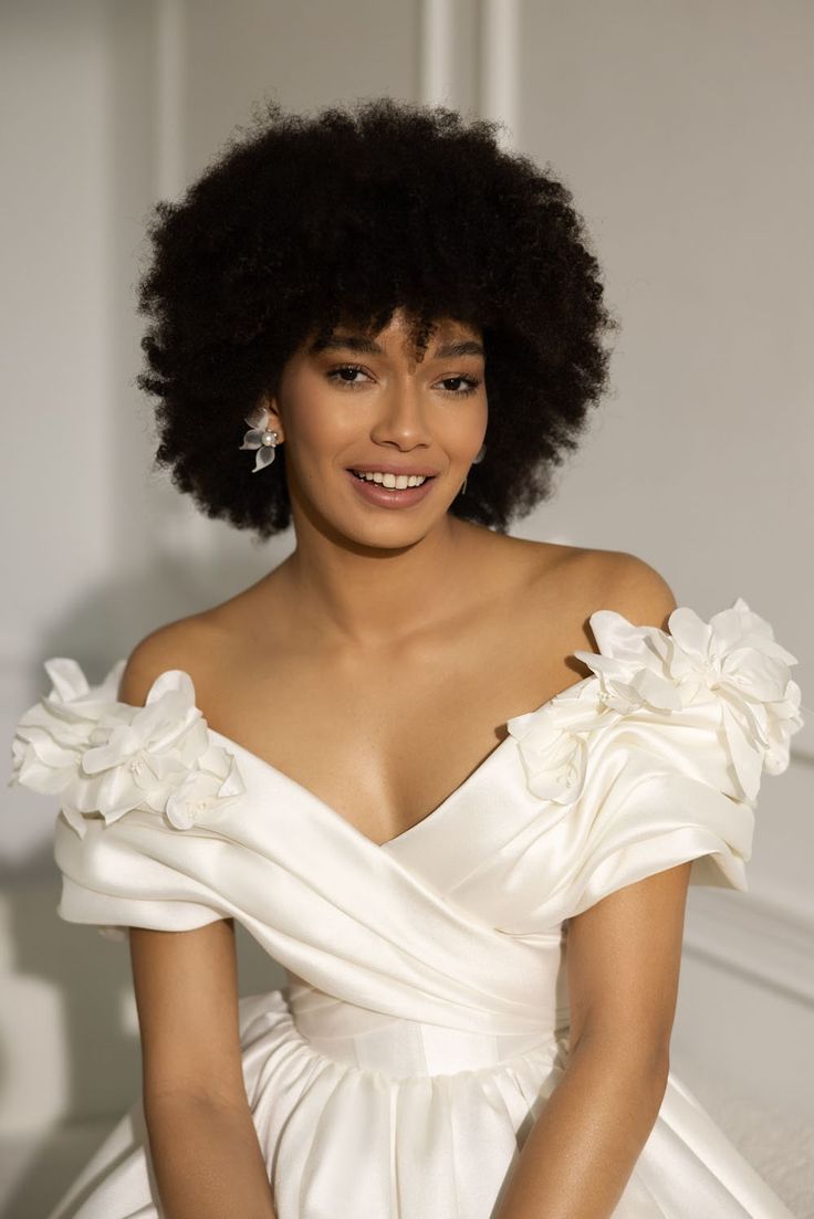 a woman with an afro in a white dress sitting on a bed and smiling at the camera