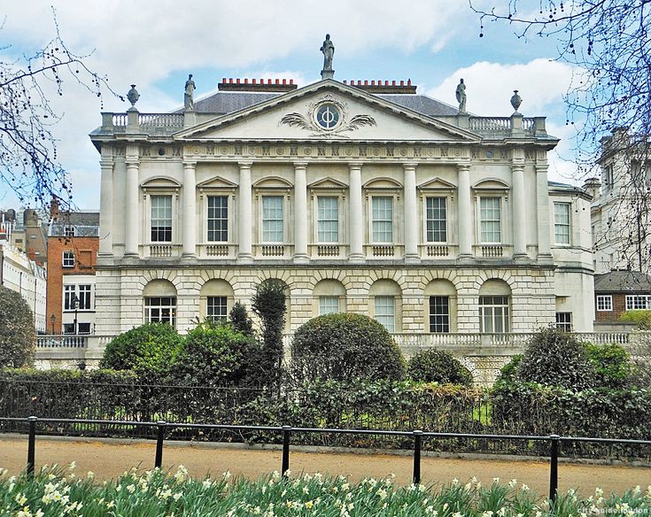 a large white building sitting next to a lush green park filled with trees and flowers