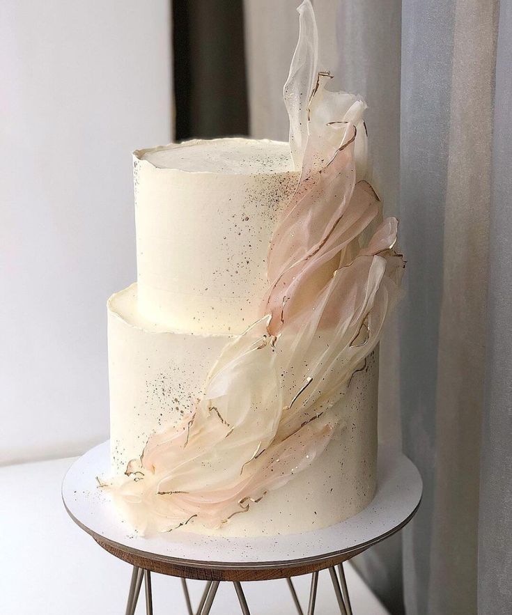 a three layer white cake with pink icing on a metal stand next to a curtain