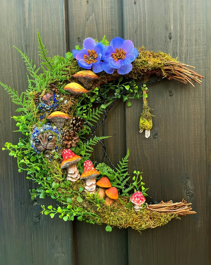 a wreath made out of moss with blue flowers and mushrooms on the front, hanging from a wooden fence