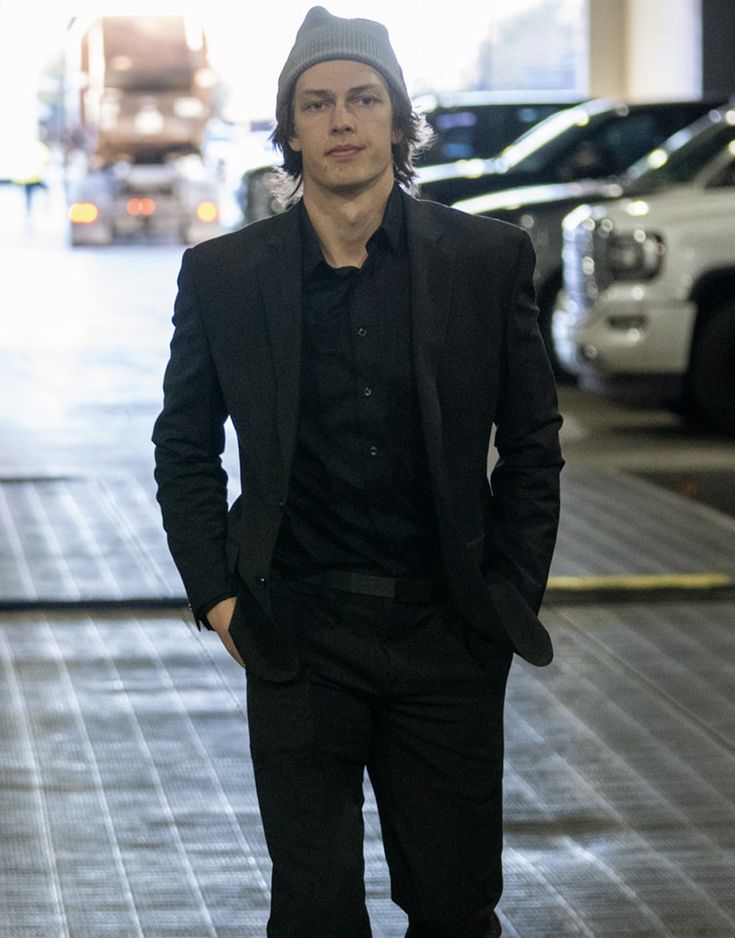 a man in a suit and beanie standing on the street with his hands in his pockets