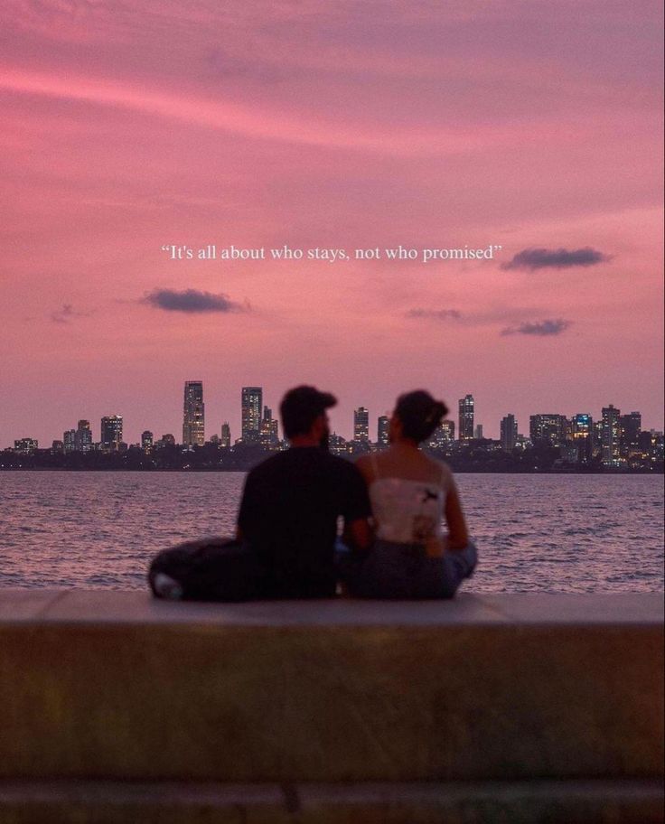 two people sitting on the edge of a pier looking at the water and buildings in the distance