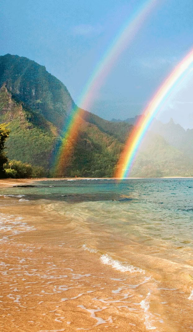 two rainbows in the sky over water and beach with mountains behind them on a sunny day