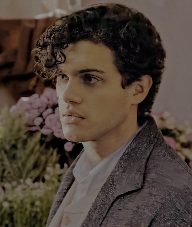 a man with curly hair is looking at the camera while standing in front of flowers