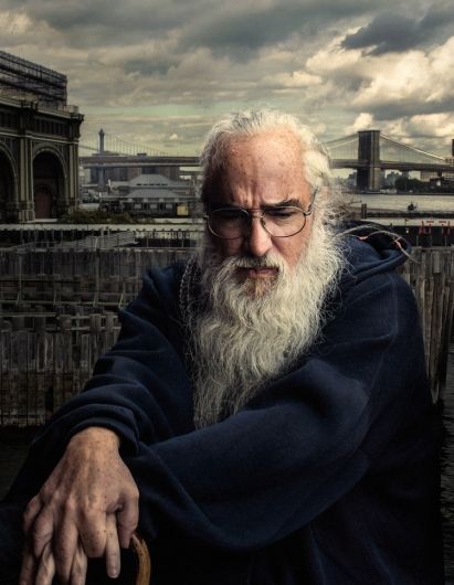 an old man with long white hair and beard sitting on a chair in front of a bridge