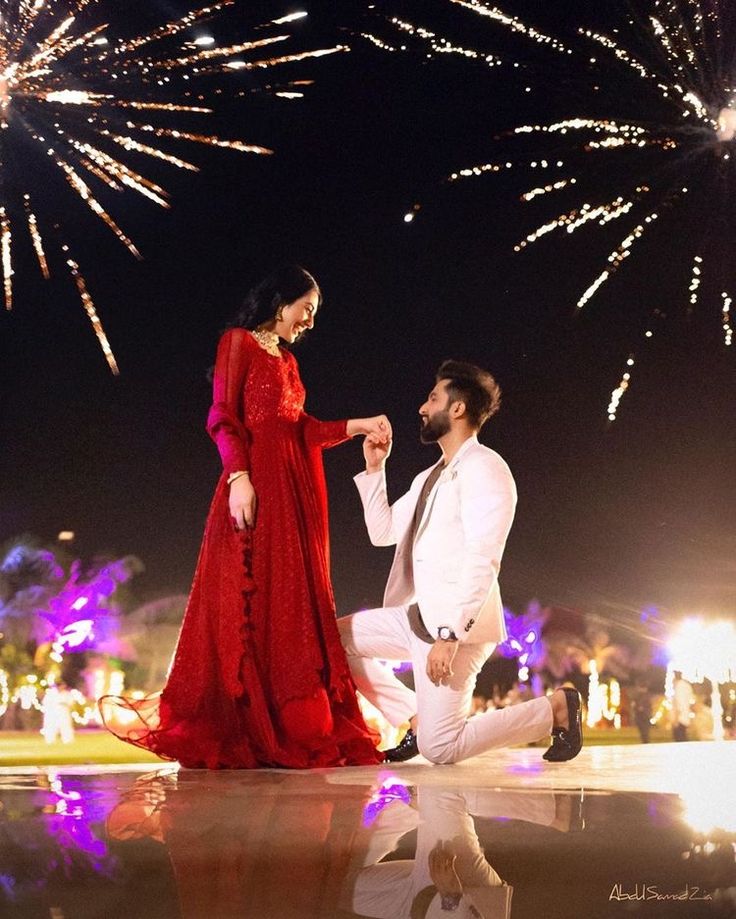 a man kneeling down next to a woman in a red dress and white suit while fireworks go off behind them