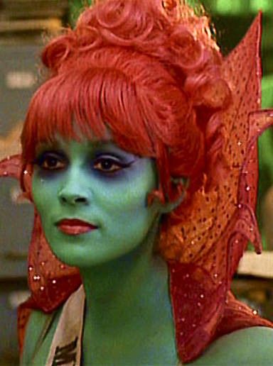 a woman with red hair and green makeup looks at the camera while sitting in an office