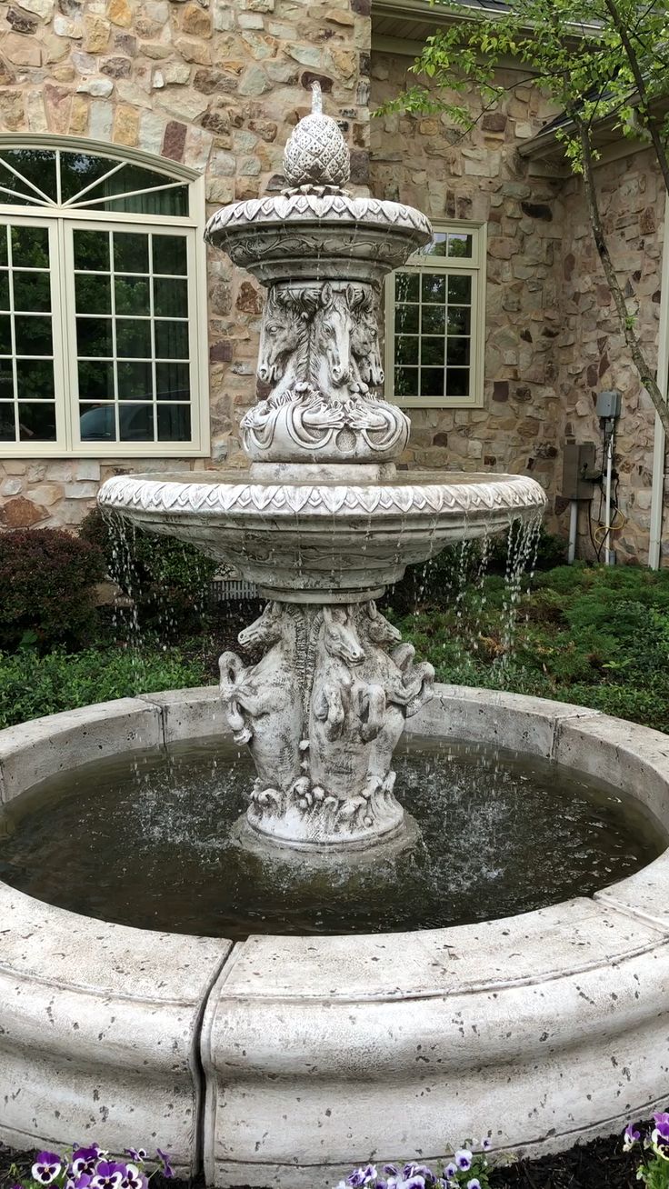 a water fountain in front of a stone building with windows and flowers around the perimeter